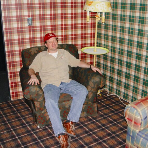 Man sits in stuffed arm chair in a set with different plaid wallpapers and tartan floor