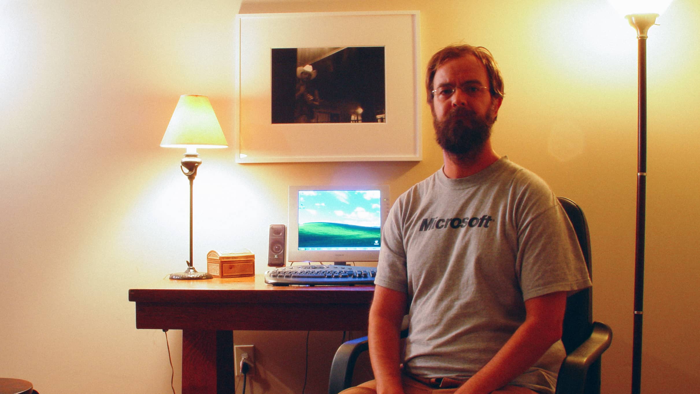 A bearded man wearing a Microsoft shirt in front of a computer running Windows