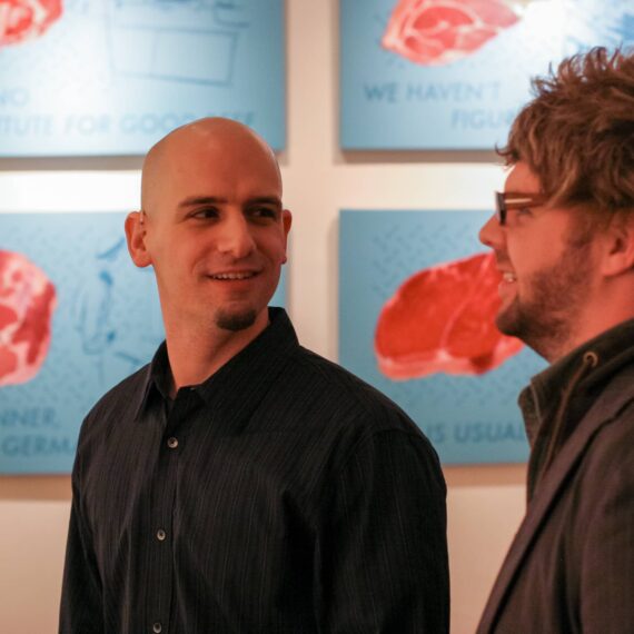 Two men at art opening with paintings featuring steaks