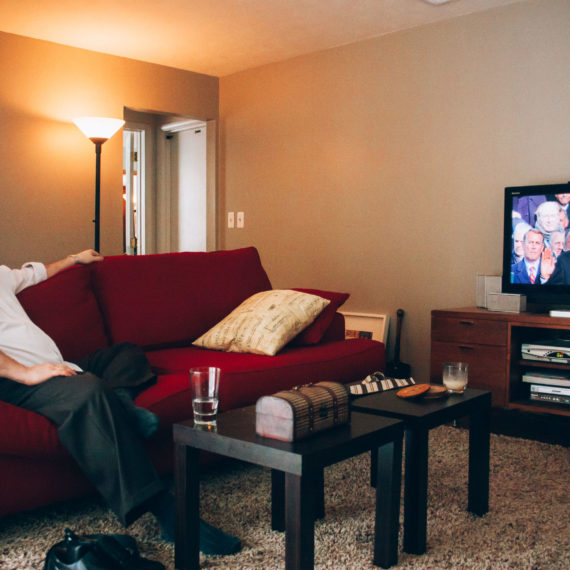 A bearded man in a dress shirt and slacks sits on a red couch and watches a television with the swearing in of President Barack Obama