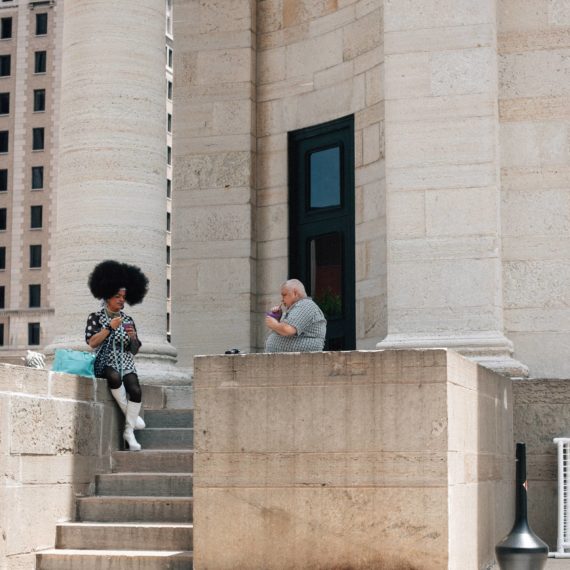 A drag queen and a man have drinks on the steps of what looks to be a courthouse, but honestly? Who knows!