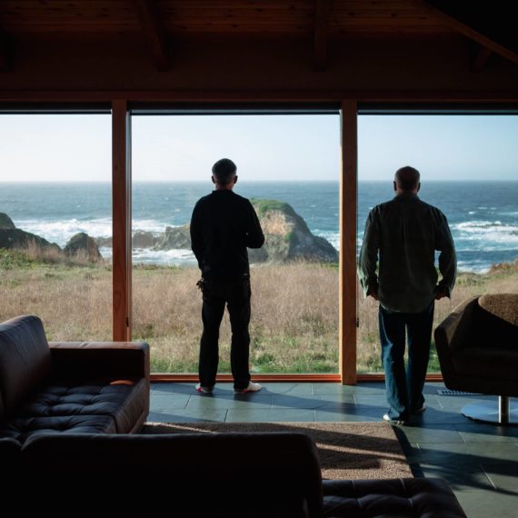 Two men look out from floor to ceiling windows in a mid century modern living room out to the sea