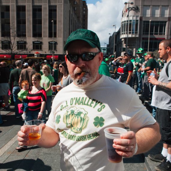 A bearded man with cigar holds two beers at a St.Patrick’s Day festivity