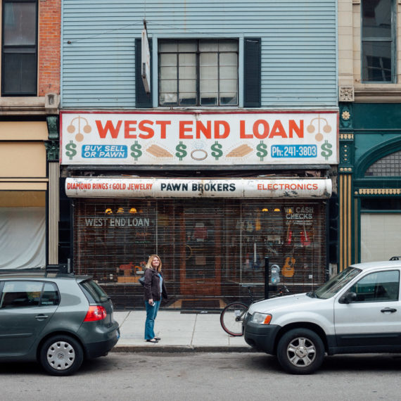 A woman stands between two parked cars on the street in front of a store with a a sign that reads "West End Loan, Buy, Sell or Pawn"