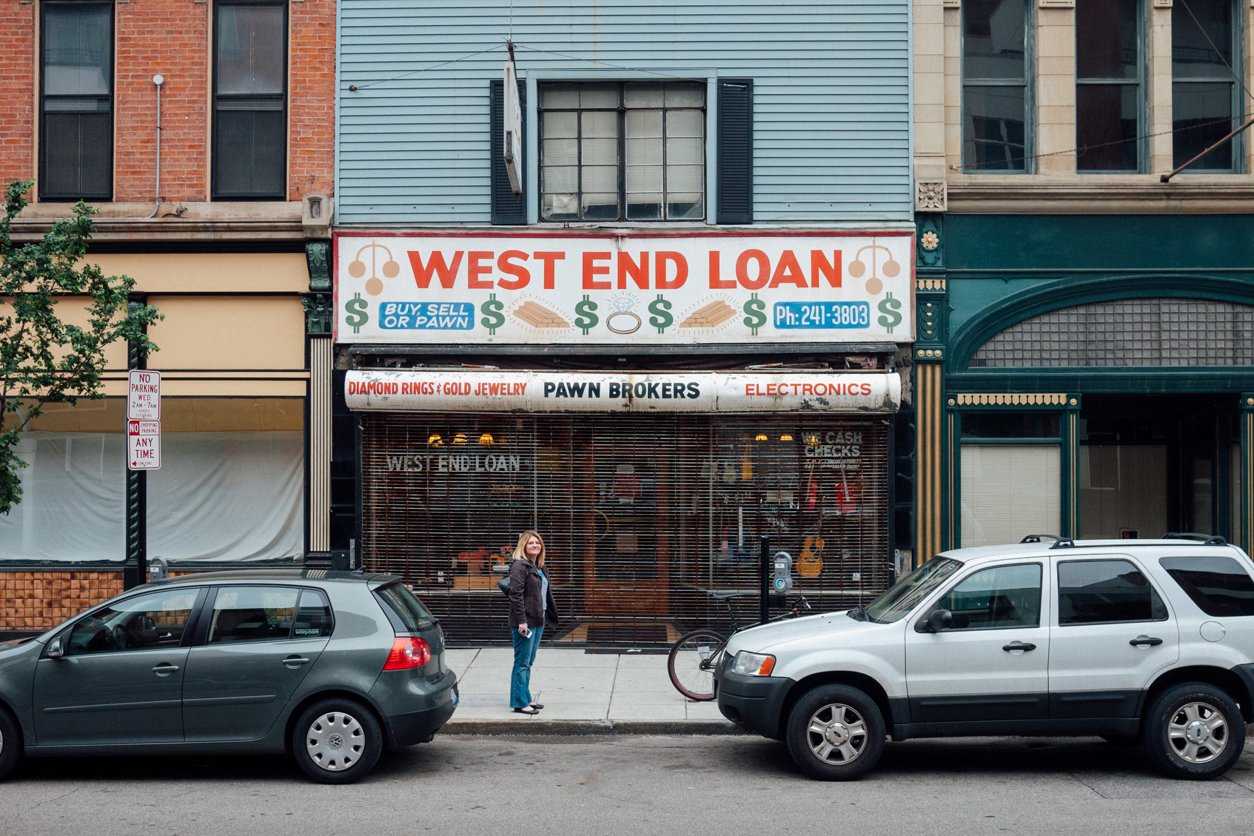 A woman stands between two parked cars on the street in front of a store with a a sign that reads "West End Loan, Buy, Sell or Pawn"