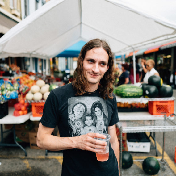 A man with long hair stands at an outside market with a drink in his hand