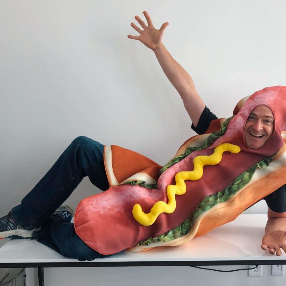 A man in a hot dog costume poses on top of a desk