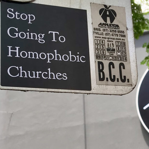 A sticker on a traffic sign that reads "Stop going to homophobic churches" set in white serif typeface on a solid black background