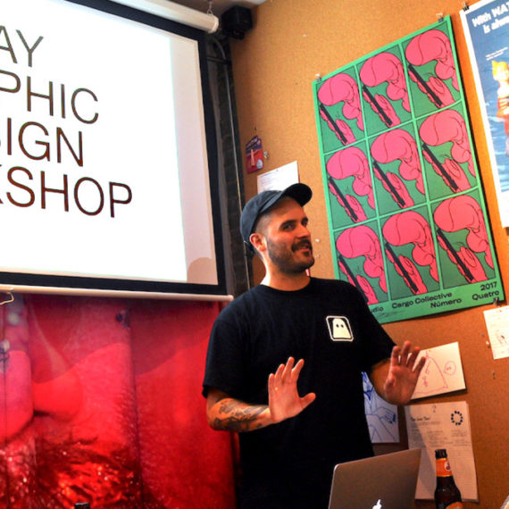 A man with stubble and a mustache wears a cap and standing in front of posters and a projected presentation with the words "Gay Graphic Design Workshop" on the screen