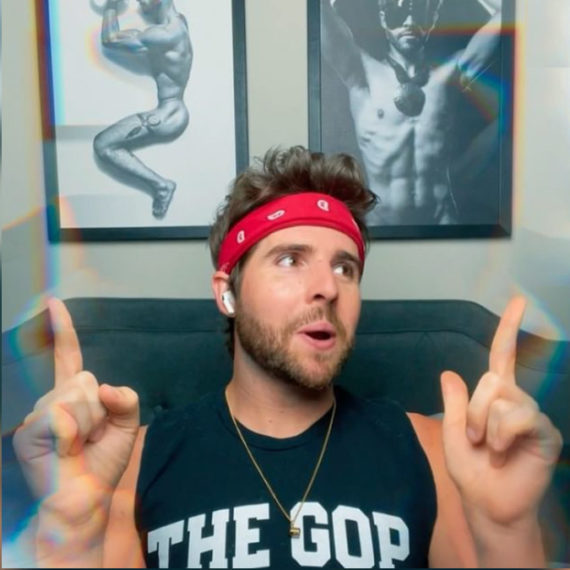 A man with a red bandana headband points both his fingers to the sky and smiles, looking off to the side. He's sitting in front of two framed black and white homoerotic art