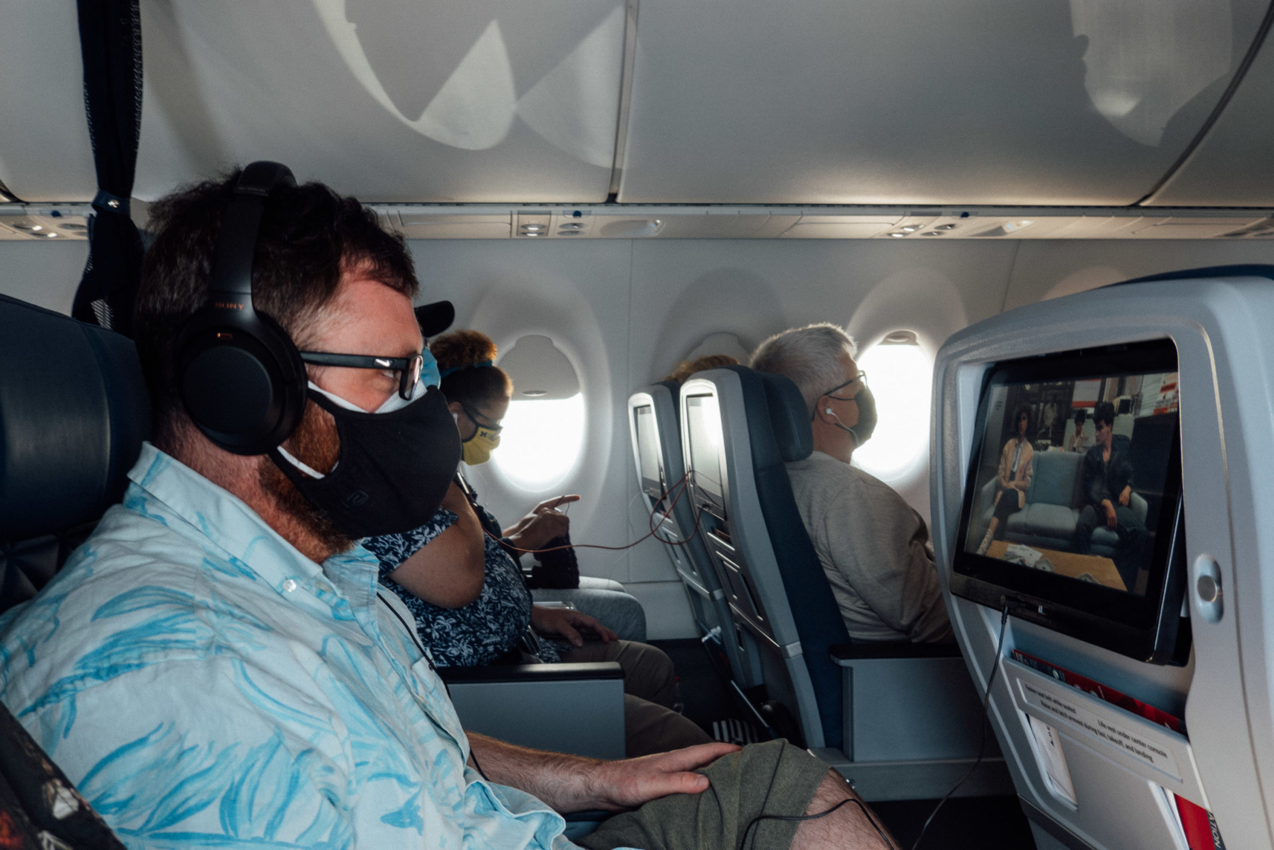 A bearded man with double facemasks on an airplane watches Ferris Bueller's Day Off on a seatback video screen