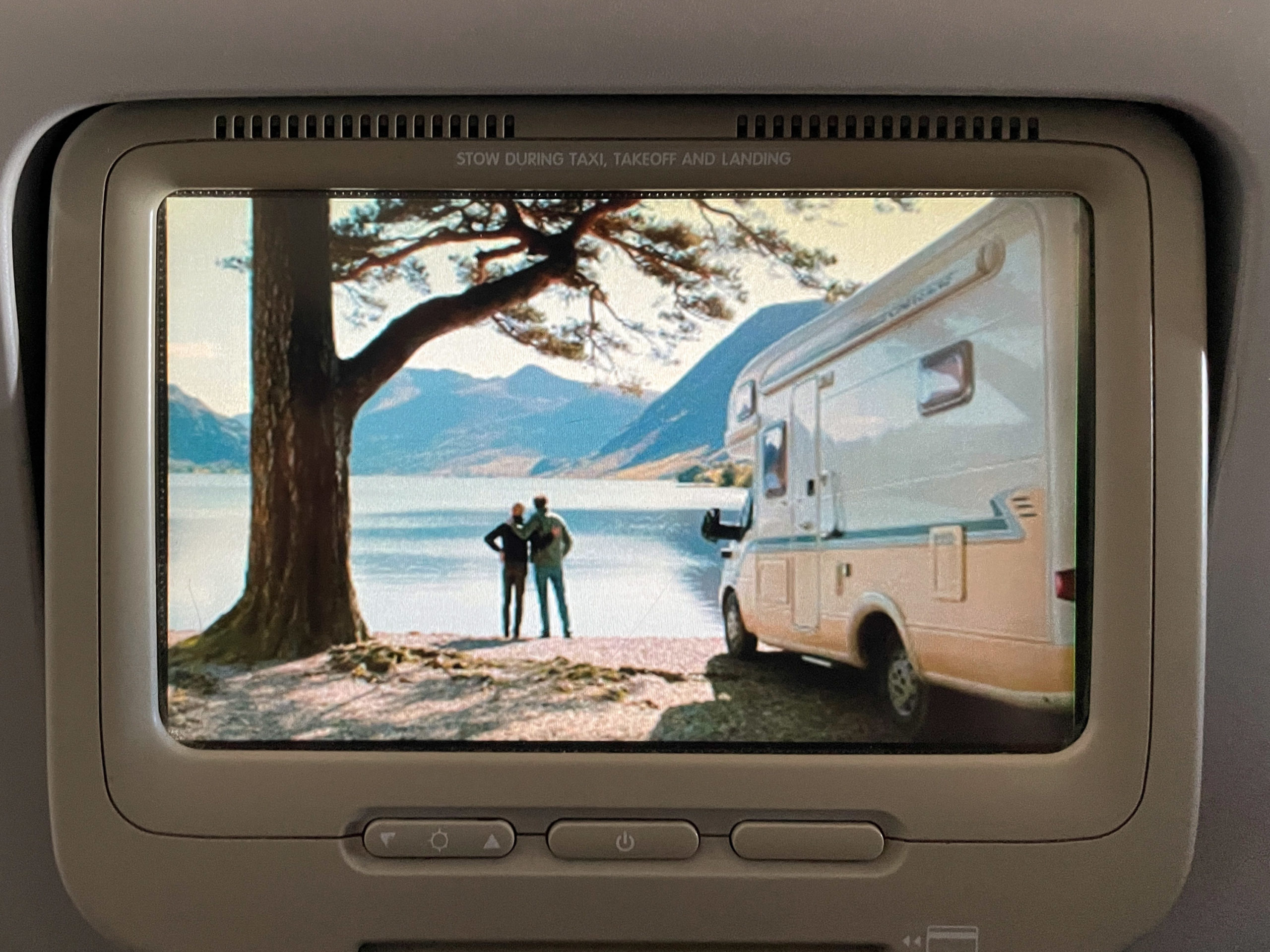 Photograph of a seatback video screen on a flight. The scene is of two men with their arms around another at the edge of a lake with an RV in the foreground