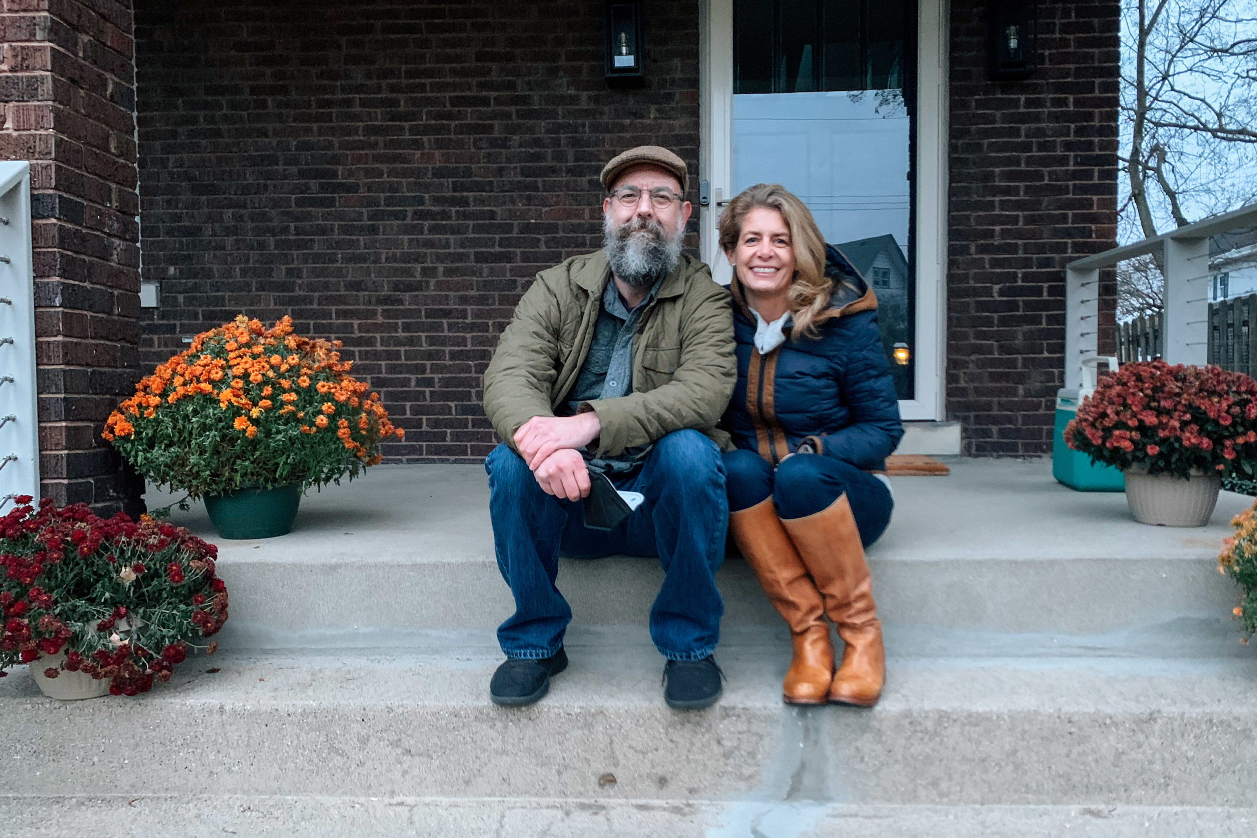 A man with a partially gray beard and glasses sits next to a woman with blond hair and tan boots on the steps of a house porch