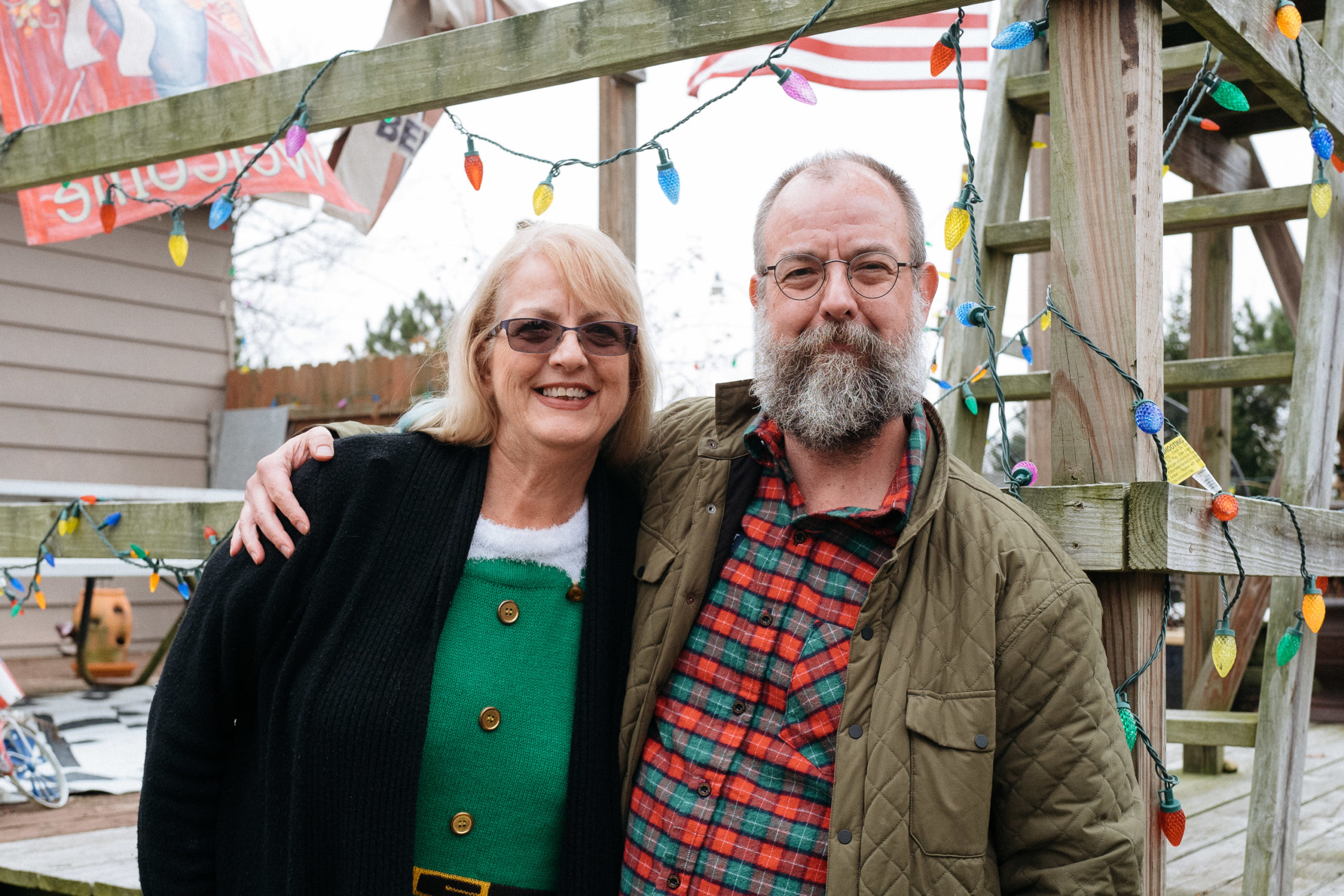 A woman in a green elf shirt stands next to her sibling in a plaid shirt. They're both smiling.