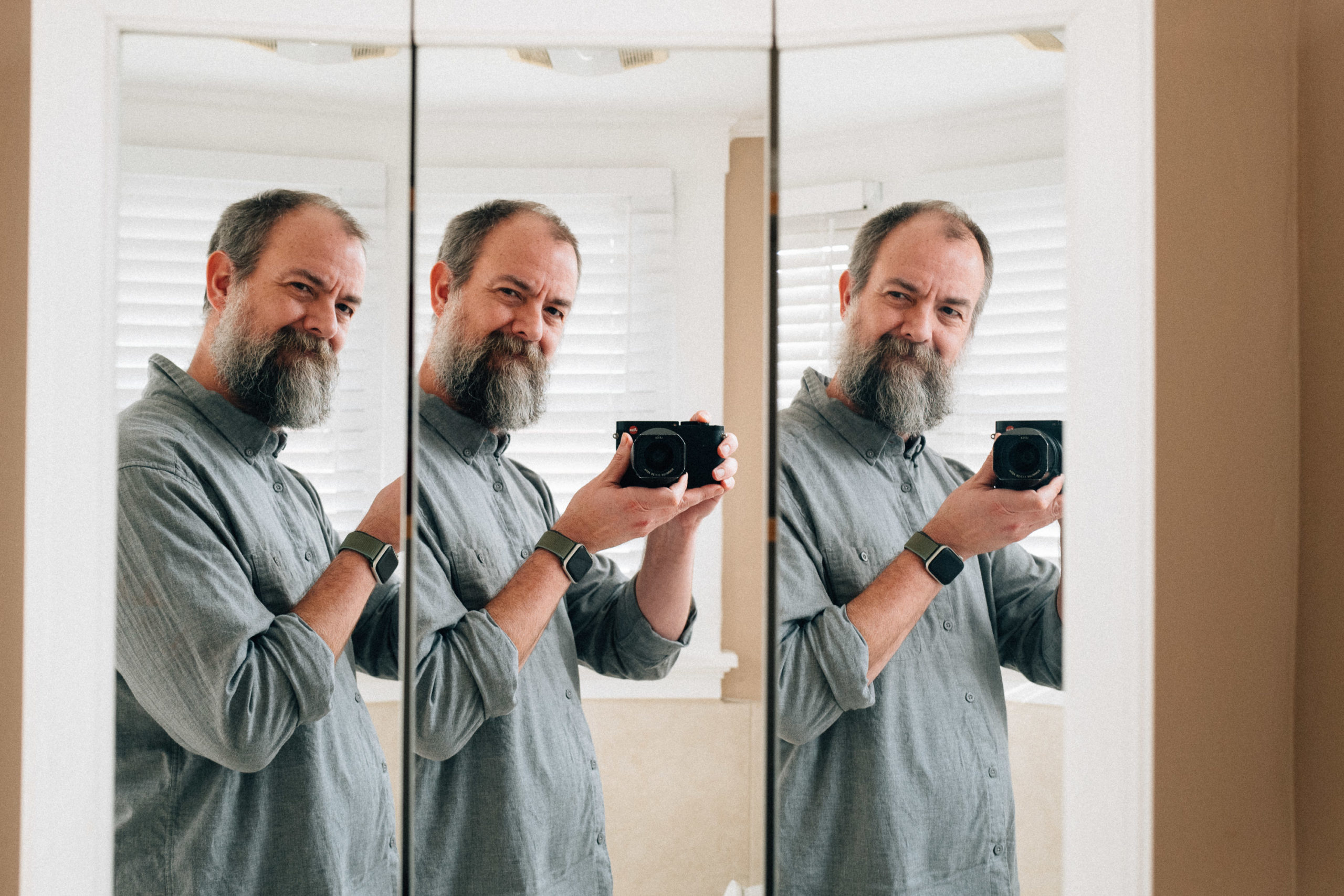 A middle aged man with a graying beard holds a camera up to a triple bathroom mirror medicine cabinet for a selfie