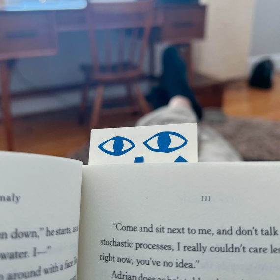 A bookmark with eyes screen printed along the top peek out of a paperback book