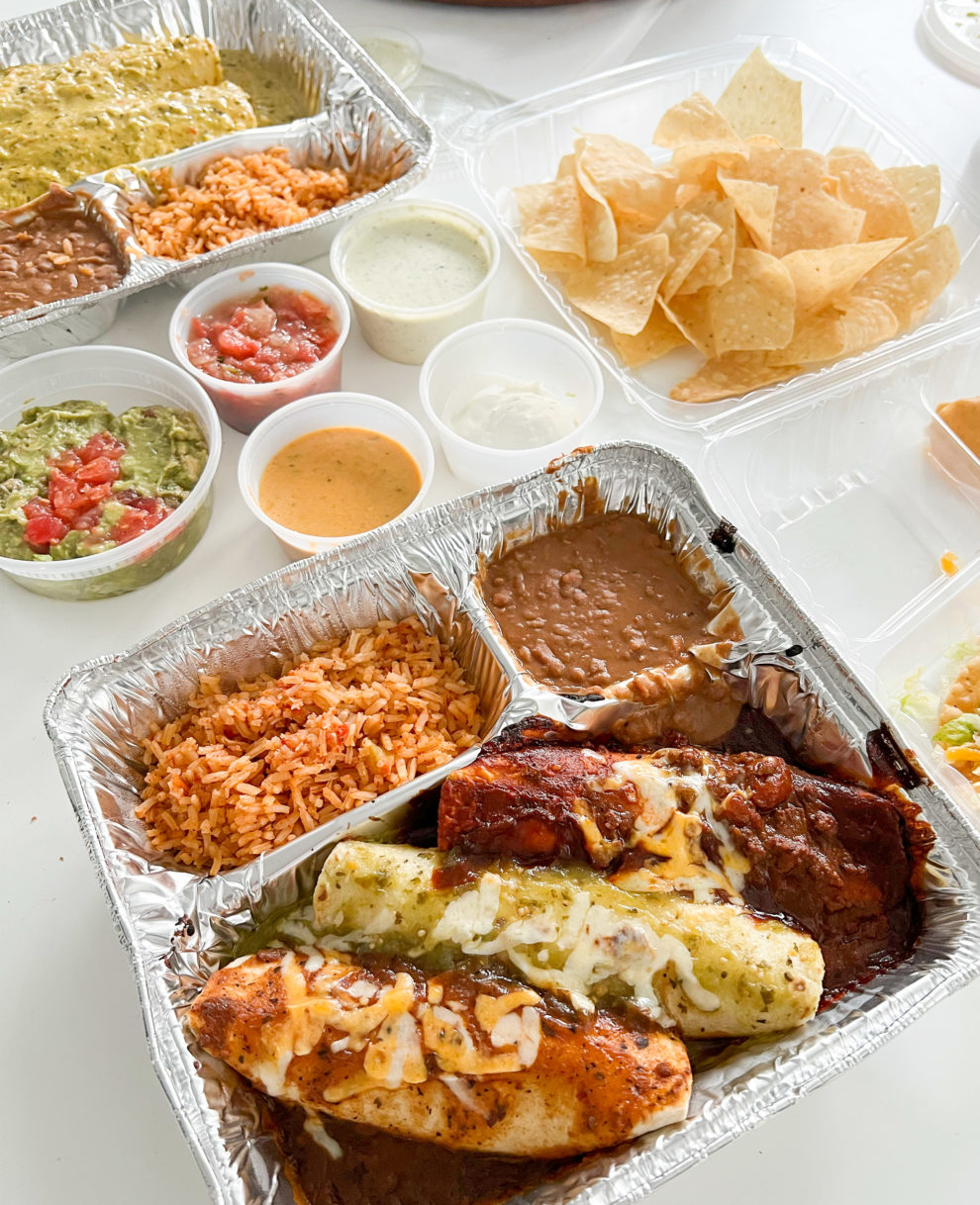 Takeout Mexican food with enchiladas, various salsas, a container of guacamole and some tortilla chips