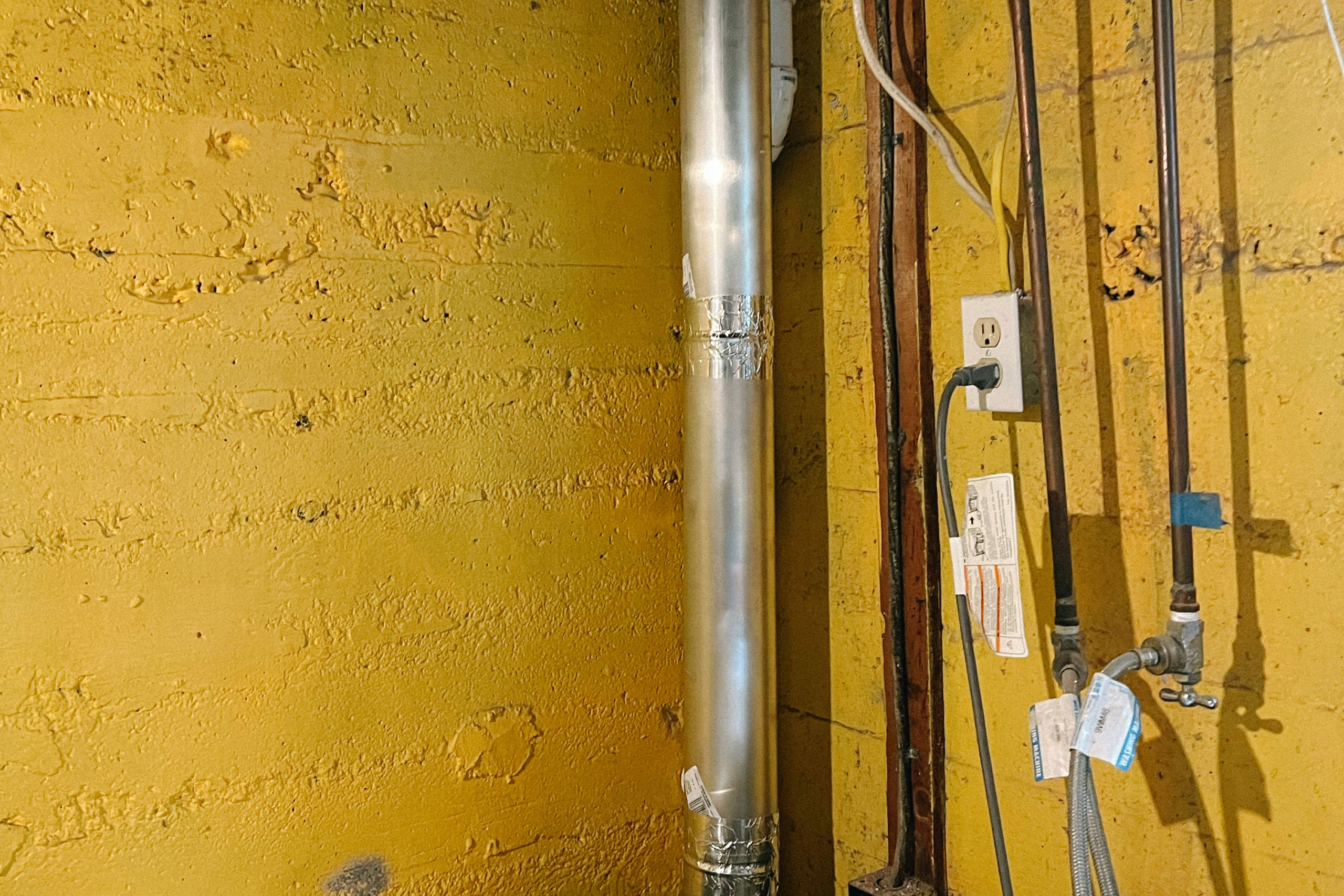 A rough textured yellow wall with metal vent, copper pipes and an electrical outlet