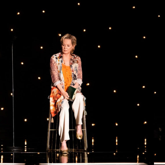 A women sits on a stool with a microphone. She's looking down toward the ground in a reflective moment or beat. She's well lit on a stage with sparkling lights on a black background that look like stars.