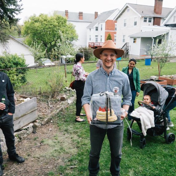 A man in a wide brimmed hat holds a covered cake at an outdoor party