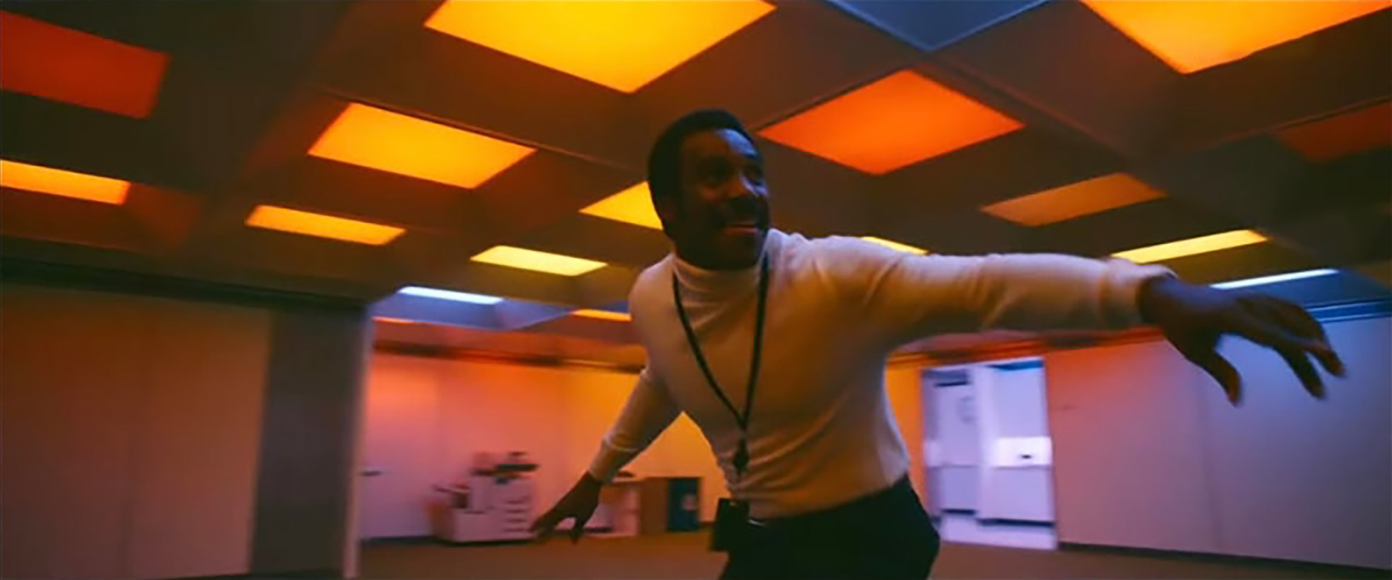 A man in a turtleneck dances in an office that is lit up like a disco