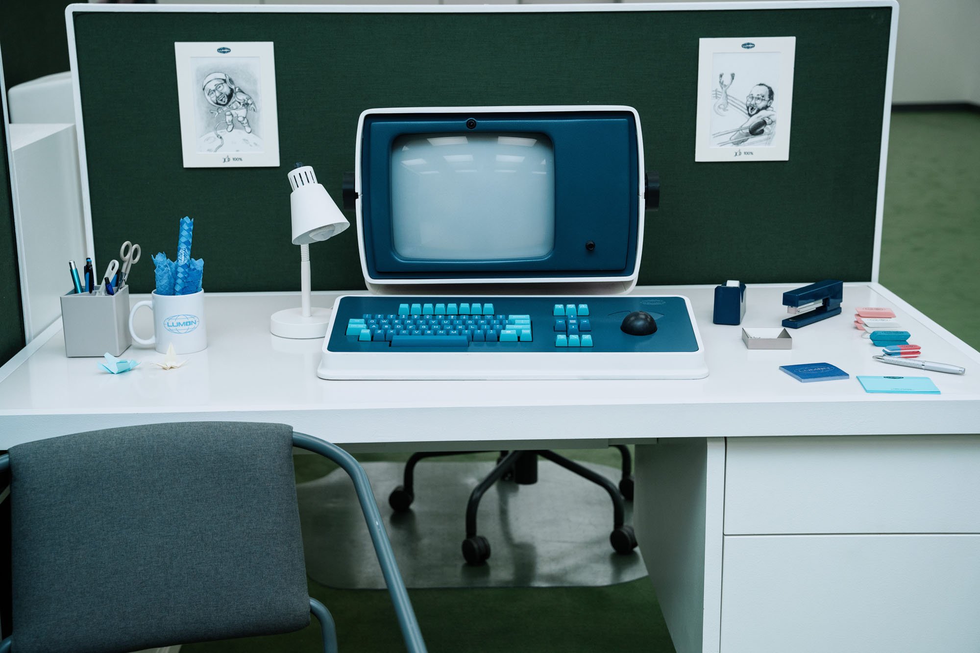 A retro computer workstation at an open cubicle with a lamp and two drawings hung on the wall