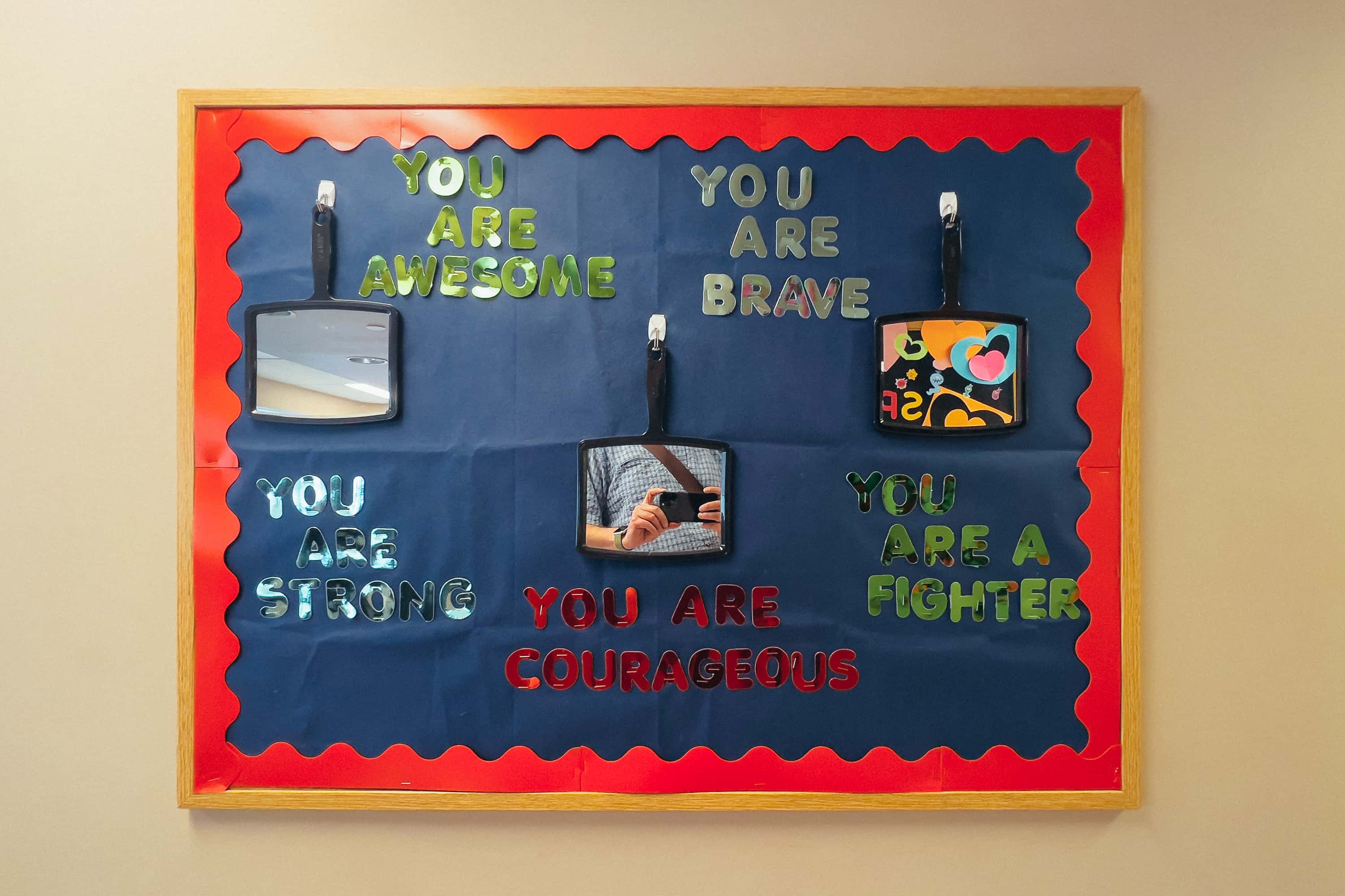 A bulletin board with supportive phrases stapled on with reflective letters that read "You are brave" "You are a fighter" "You are awesome." with three mirrors on hooks