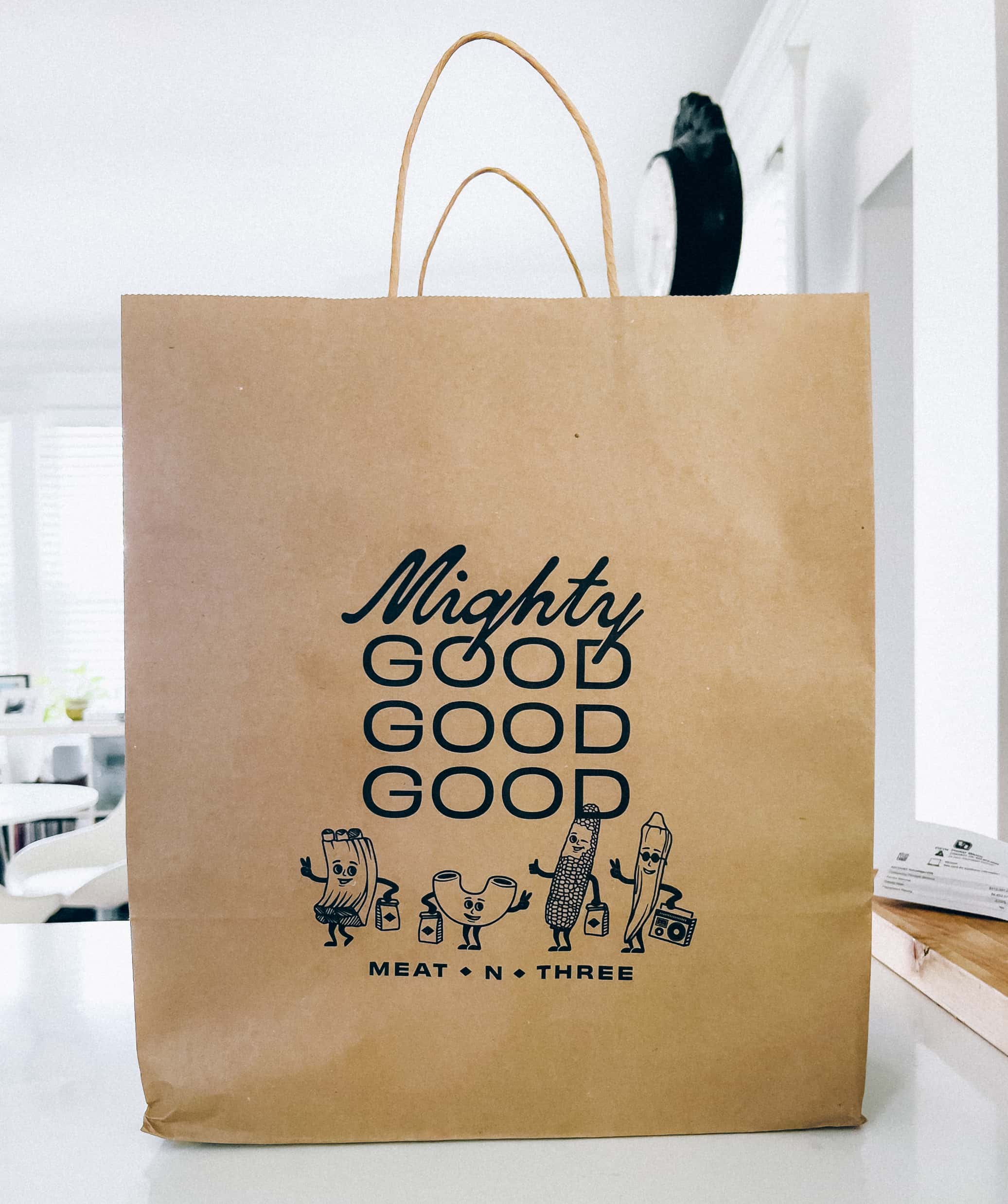 A brown bag with handles that has a stamp design that says Mighty Good Good Good with anthropomorphic cartoons of food items along the bottom