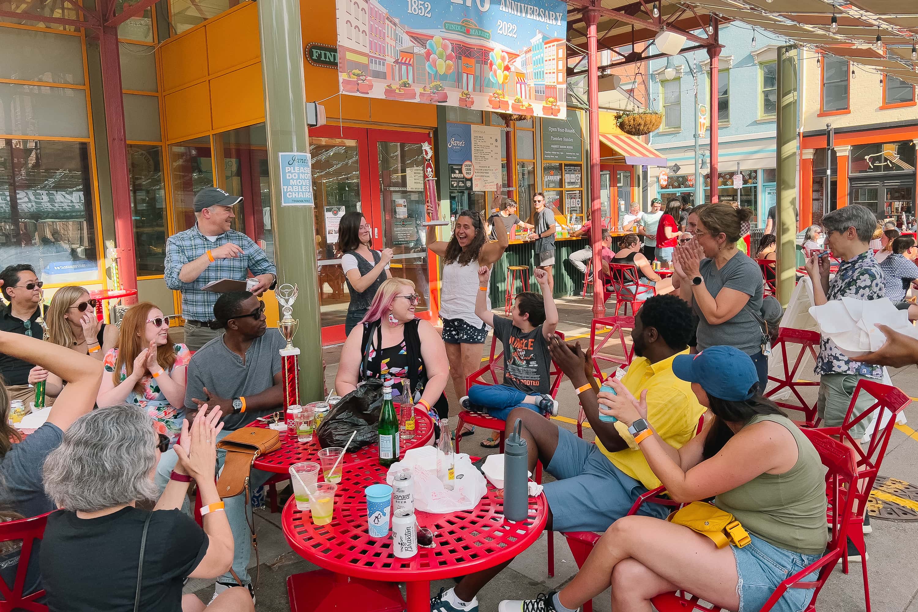 A group of folks at an outdoor patio bar of a farmer's market on a sunny afternoon. Their attention is directed at a woman holding a large trophy over her head.