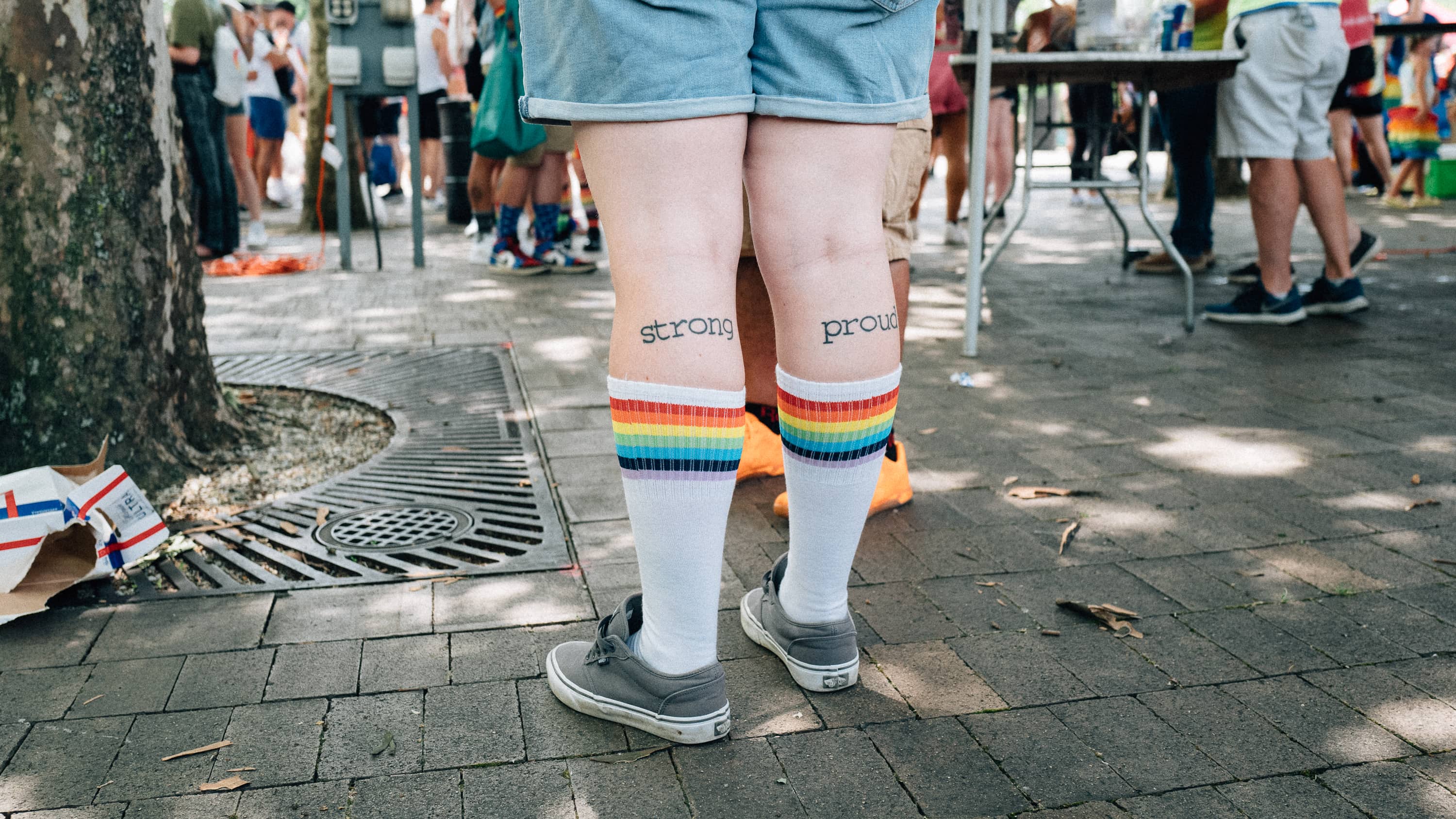 A person has rainbow striped socks and tattoos that read "strong" on the left calf and "proud" on the right