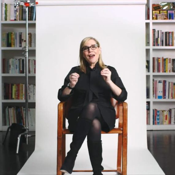 A woman sits in a chair on a white rolling backdrop of paper in front of a library of books