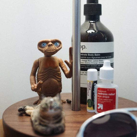 A toy action figure of E.T. is on the base of a desk lamp with a bottle of lotion, chap stick, eye drops, a small sculpture of a cat and sunglasses