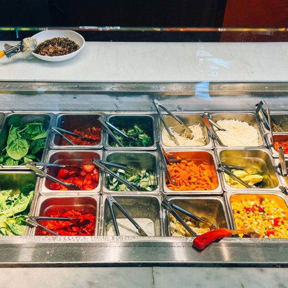 Ingredients at a fast casual restaurant that include lettuce, grains, peppers, carrots, corn, pickled onions, hummus, broccoli, cheese, strawberries, olives, cabbage and avocado