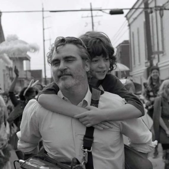 Black and white still from a movie where a man in a white button down shirt carries a young man on his back in the midst of a parade in New Orleans