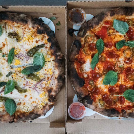 Two takeout pizzas. One with olive oil, corn, shishito, and chili crisp and the other with pepperoni and basil