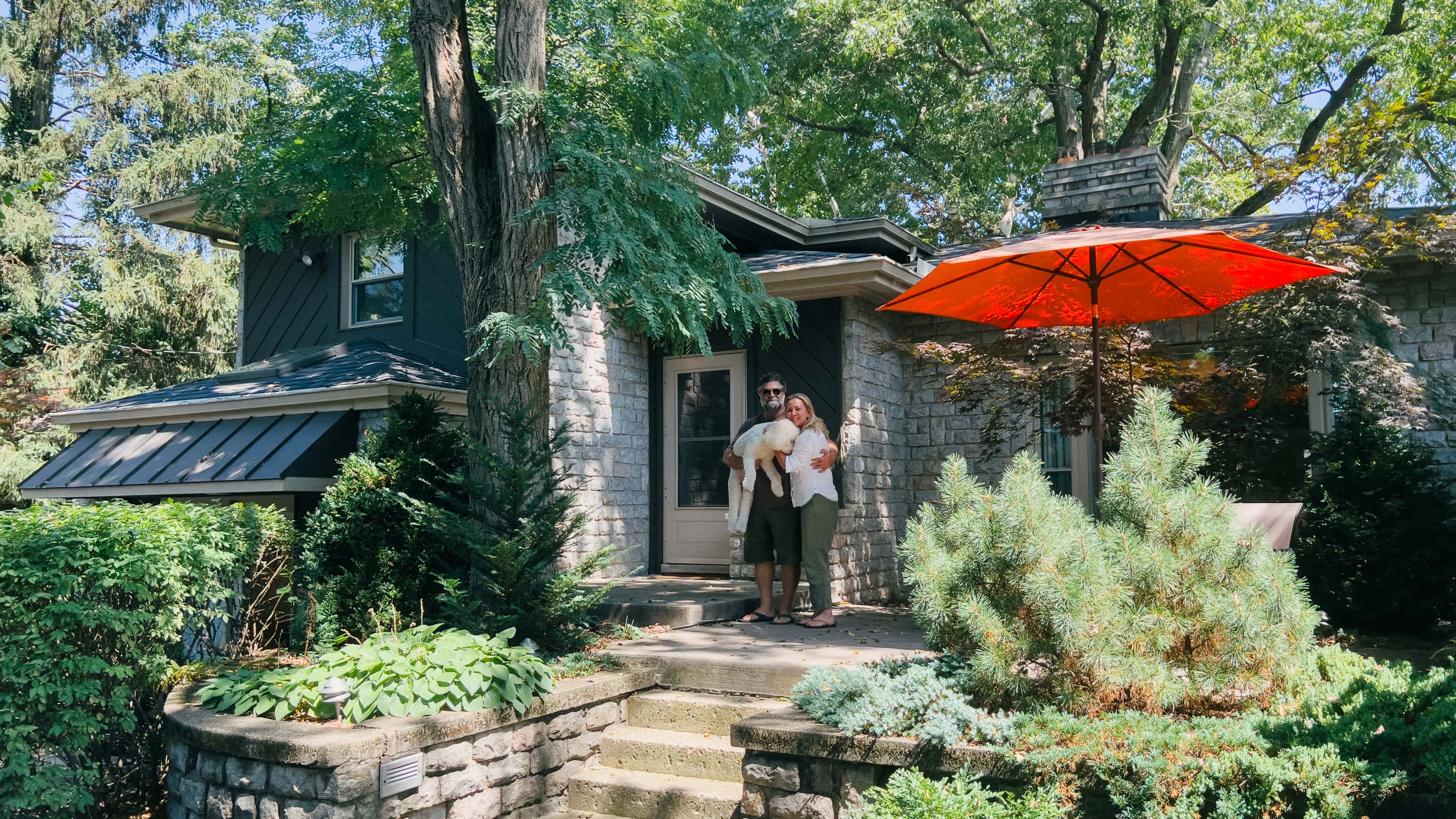 A man and woman stand in front of a house holding a medium sized fluffy white dog in their arms. The dog is being shy