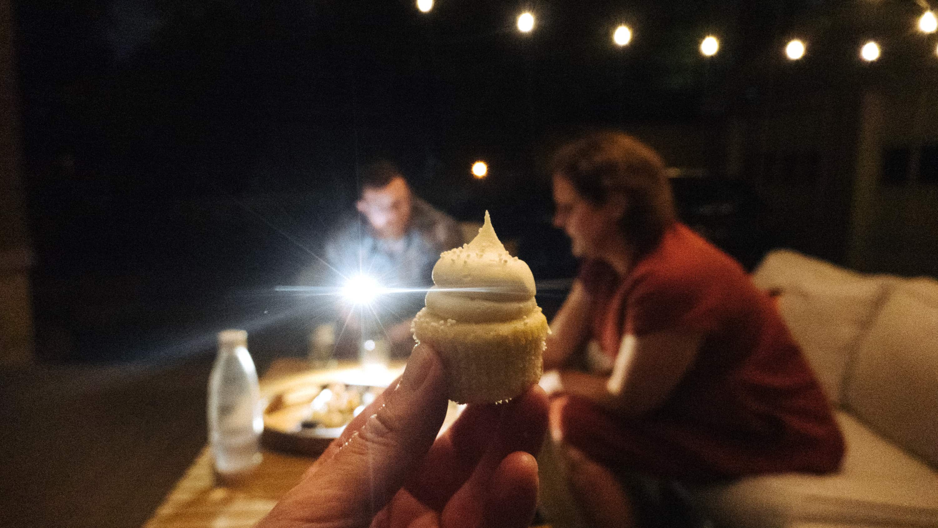 A person holds a mini cupcake in front of the camera at night while two figure in the background search through a box of remaining cupcake flavors