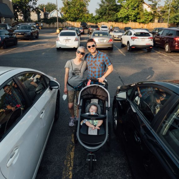 Melissa & Kyle stand with their child in a stroller in the parking lot of a grocery store