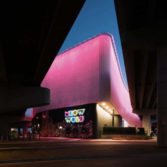 An angular building lit with pink lights framed by the shadows of highways. The base of the building has a neon sign that reads MEOW WOLF