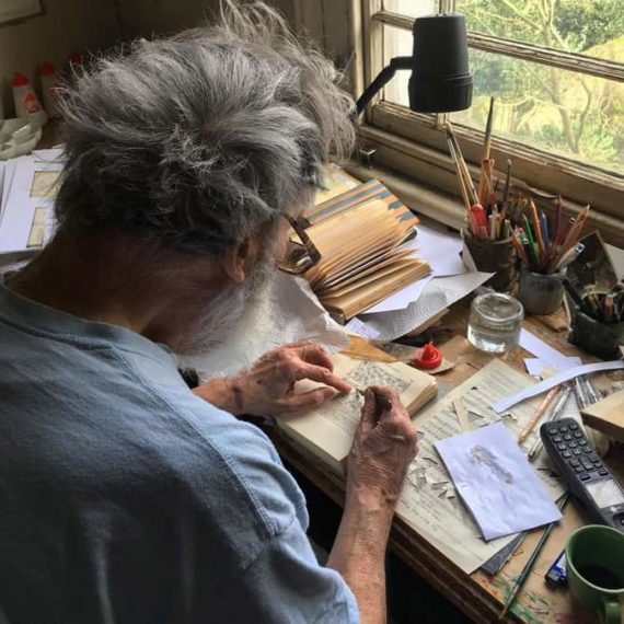 An older fella works at a desk filled with instruments for making art