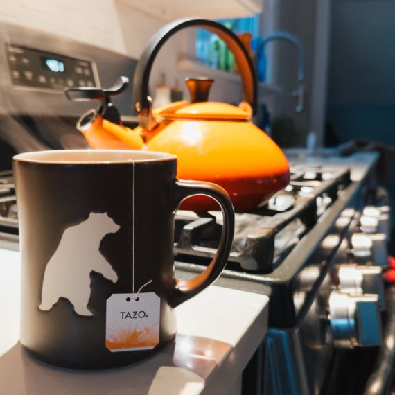 A mug of tea steeping on the counter next to a stove with an orange teapot