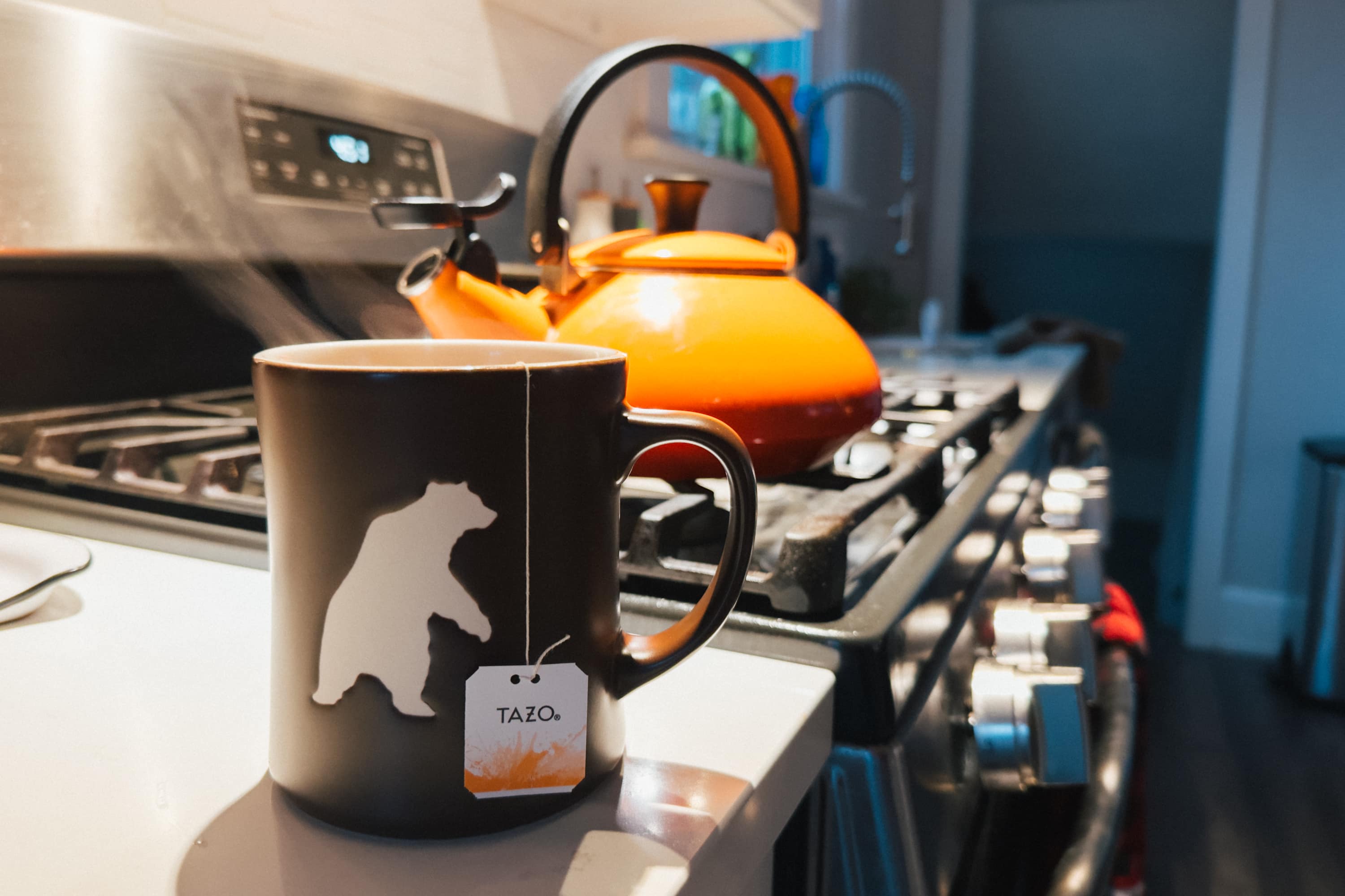 A mug of tea steeping on the counter next to a stove with an orange teapot