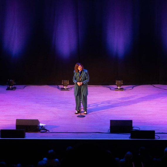 Paula Pondstone on stage in a striped suit and purple lights