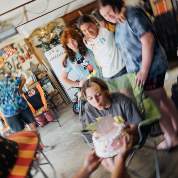 Blowing out candle on a birthday cake, surrounded by friends and family in a garage