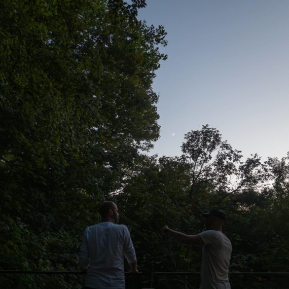 Two men stand on a porch looking out over the trees