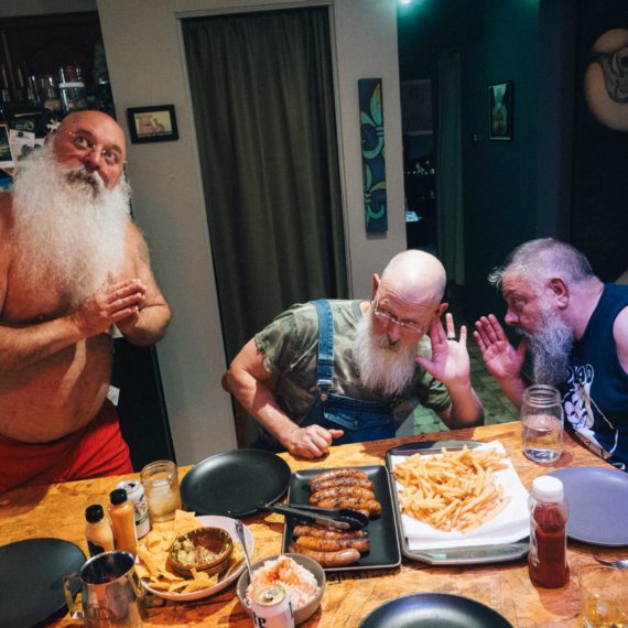 Three bearded men around a dinner table filled with sausages and French fries