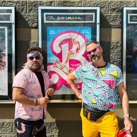 Men dressed in day-go 80s stand in front of a Barbie movie poster 2023