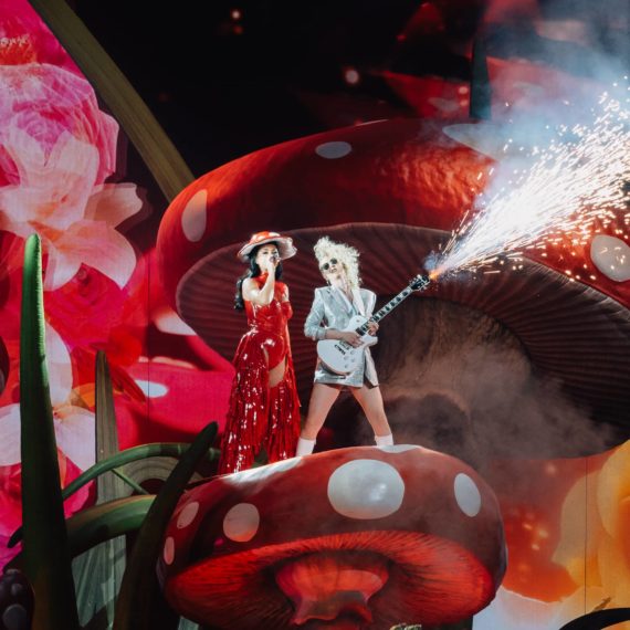 Katy Perry on stage in a mushroom themed costume on top a mushroom with a guitarist who has fireworks streaming from the neck of the guitar