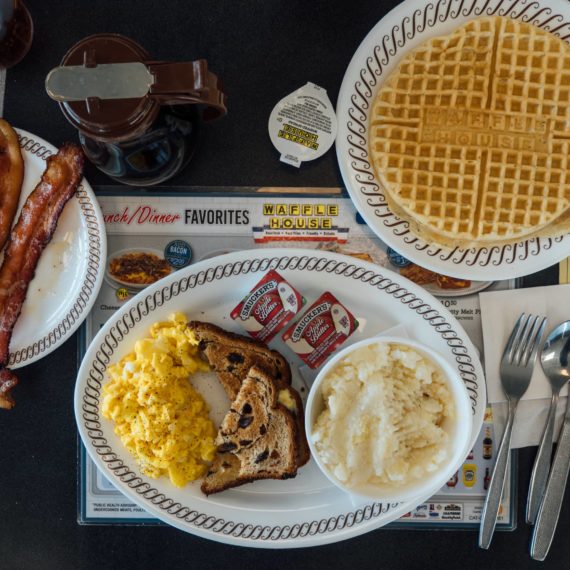 Breakfast at Waffle House that includes scrambled eggs with cheese, raisin toast, apple butter, grits, a waffle and bacon