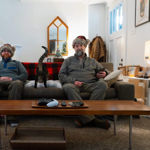 Two men in Santa hats sit on a couch with a cat